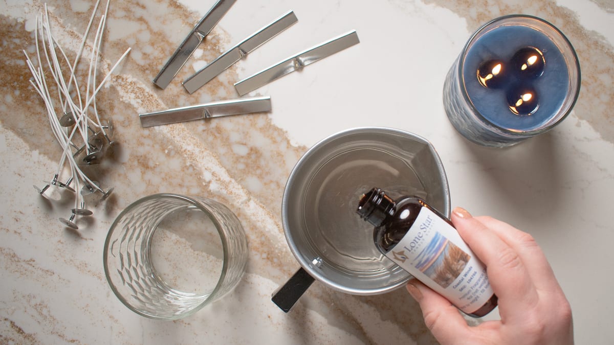 A hand pouring a bottle of Lone Star fragrance oil into a metal pouring pot, beside candle wicks, an empty glass jar, and a finished blue candle