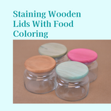 Four small glass jars with wooden lids stained tan, pink, green and mint, text reading Staining Wooden Lids With Food Coloring