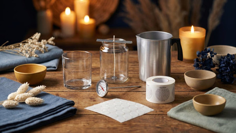 Candle-making supplies on a wood table: empty glass tumbler, wicked jar, dial thermometer, aluminum pouring pot, wick stickers, and a roll of caution labels, with lit candles glowing behind