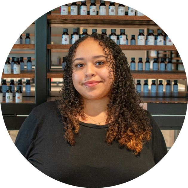 smiling woman with curly dark hair in a black top before wooden shelves of labeled bottles