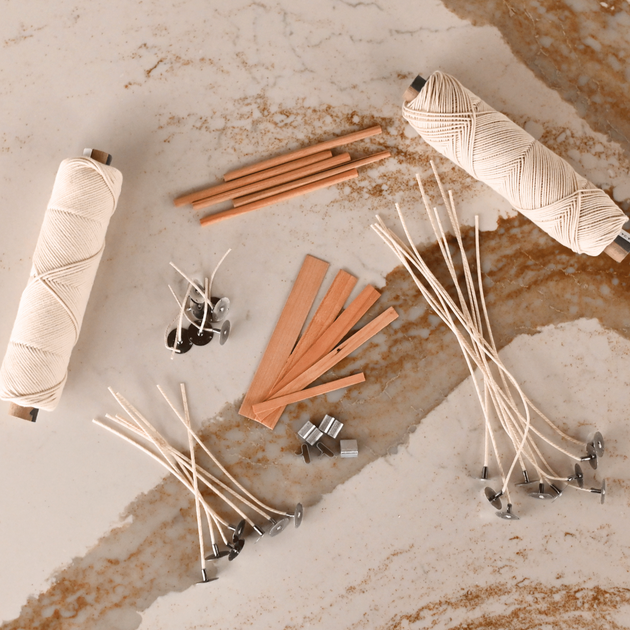 Assorted candle wicks on a marble surface: two spools of cotton wick, wooden wicks, and pre-tabbed waxed wicks with metal tabs