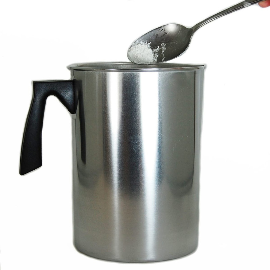 Spoon dropping white stearic acid powder into a stainless steel candle pouring pitcher