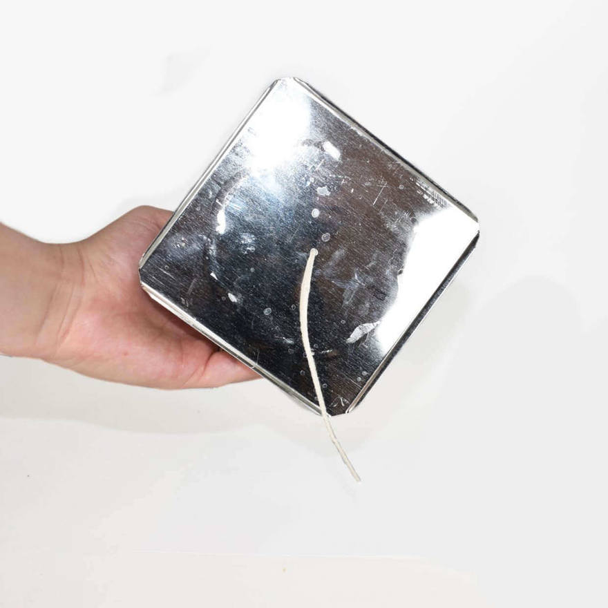 Hand holding the shiny square base of an upside-down metal pillar mold, wick threaded through