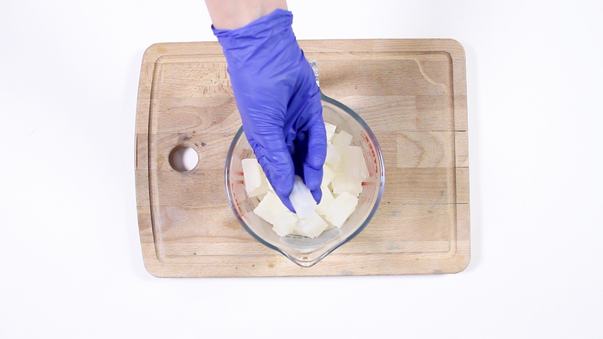 Purple-gloved hand placing white soap base cubes into a glass measuring bowl on a wooden cutting board