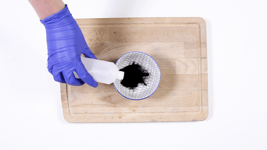 Blue-gloved hand pouring liquid from a white bottle onto black charcoal powder in a dotted bowl