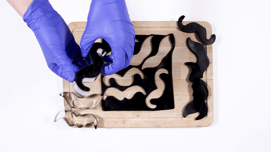 Blue-gloved hands cutting moustache shapes from black soap with metal cutters on a wooden board