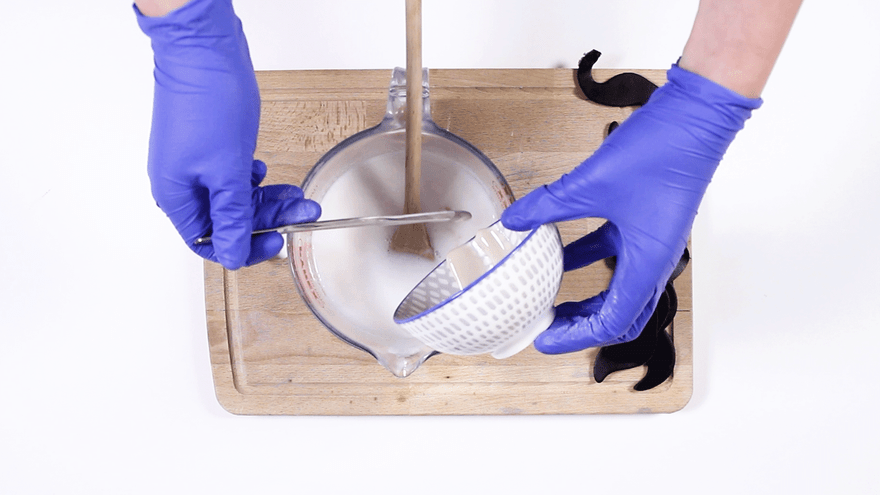 Blue-gloved hands straining white melted soap through a dotted bowl into a glass jug, black moustaches alongside