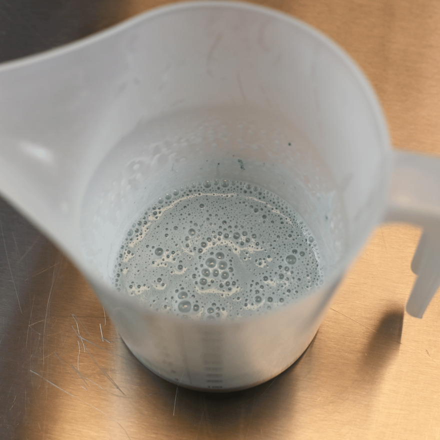 Plastic measuring jug holding melted pale blue-green soap with foamy bubbles on top