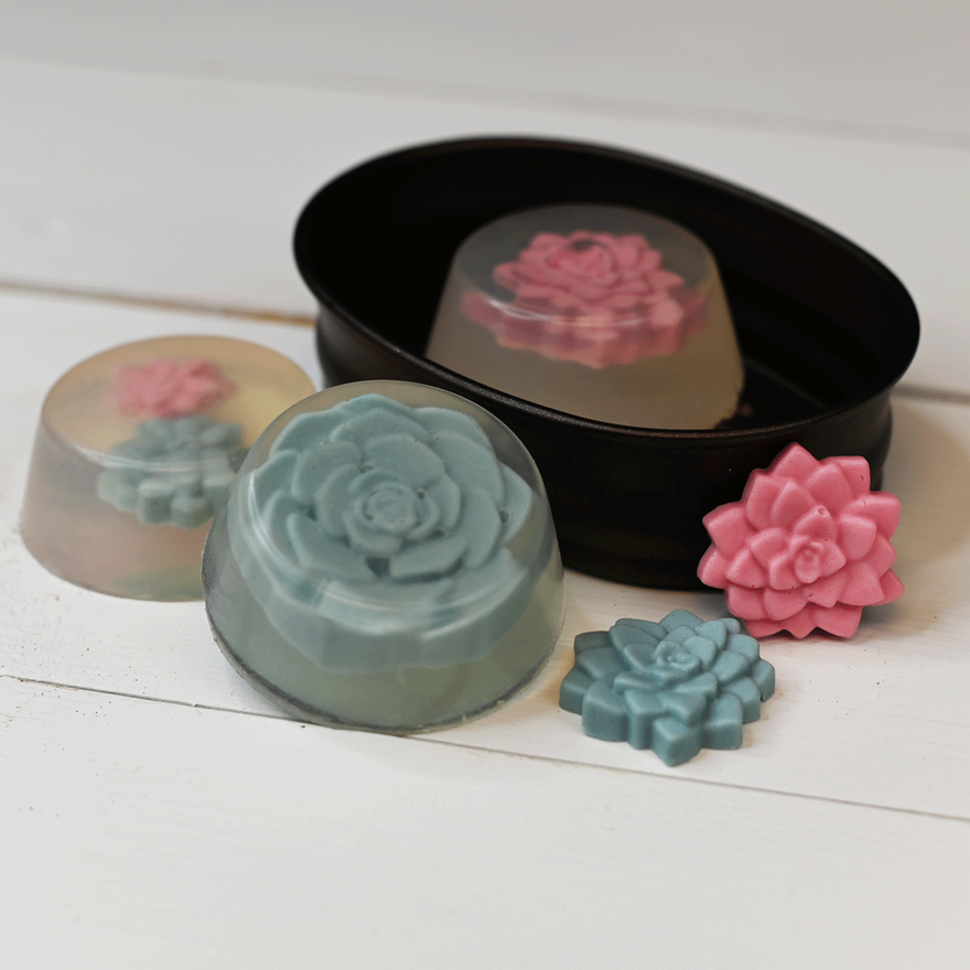 Finished blue and pink succulent soaps beside a black oval tin holding a clear soap with pink rose embed
