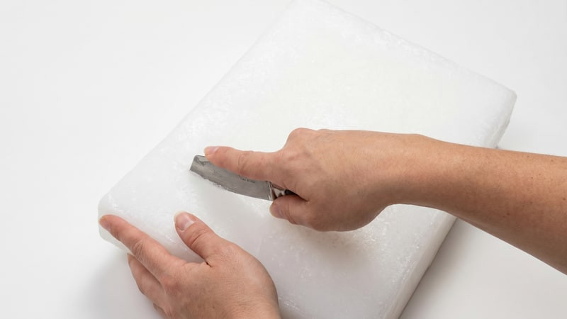 Hands using a putty knife to chip into a white slab of votive wax